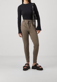 Zwarte geribbelde top met lange mouwen in combinatie met aangesloten kaki broek met een elastische tailleband. Draagt chunky zwarte sandalen met studs.