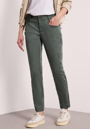 Persona che indossa pantaloni verde aderenti, giacca corta beige, t-shirt bianca e scarpe da ginnastica bianche, in piedi su pavimento grigio.