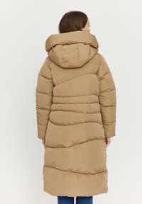 Long camel-colored puffer coat with a hood, featuring quilted patterns and horizontal seams, extending to mid-calf, paired with blue jeans.