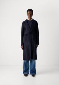 Mulher em pé, vestindo um longo casaco preto com cinto, suéter azul-marinho, gola de camisa bege, jeans largos azuis e sapatos pretos, contra um fundo branco liso.