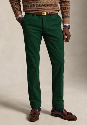 Homme portant un pantalon vert foncé, une ceinture marron, un pull à motifs, des mocassins marron à glands et une montre-bracelet, debout les mains dans les poches.