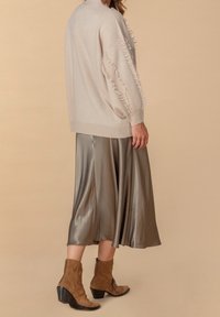 Beige gestrickter Pullover mit Fransendetails an den Ärmeln, kombiniert mit einem fließenden satinschicken oliven Rock und braunen Wildleder-Stiefeletten mit Absatz.