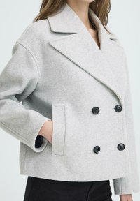 Femme portant un manteau en laine gris clair à double boutonnage avec des boutons noirs et une main dans la poche, associé à un pantalon noir.