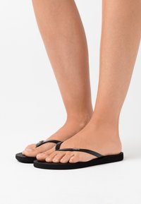 Des tongs noires avec une semelle texturée et des fines brides. Présentent un petit accent de logo métallique sur la bride. Conçues pour un usage décontracté.