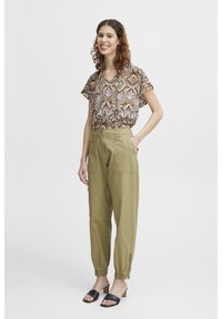 Haut à manches courtes marron à motif avec un col en V ; pantalon fuselé vert olive clair avec poches ; sandales à talons blocs ouvertes noires.