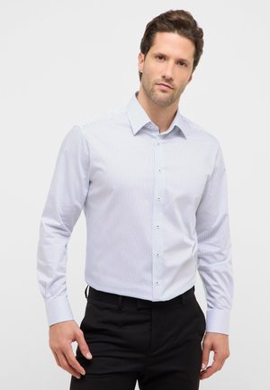 Man draagt een wit overhemd met subtiel zwart stippenpatroon en zwarte broek, staand tegen een effen lichte achtergrond.