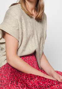 Pull en maille torsadée beige oversize associé à une jupe plissée rouge ornée d'un motif floral et de pois. Détails des vêtements texturés et superposés visibles.