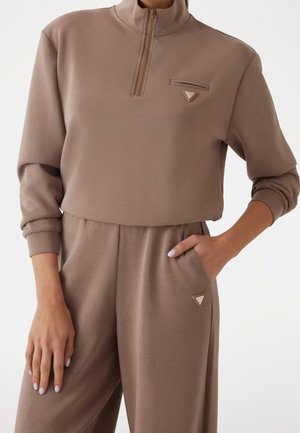 Mujer con una camiseta taupe de manga larga con cremallera hasta el cuarto de altura y pantalones a juego con detalles de logotipos metálicos triangulares, una mano en el bolsillo y la otra relajada.