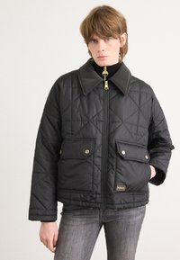 Casaco acolchoado preto com gola alta, zíper dourado e dois bolsos frontais. Apresenta costura diagonal e uma etiqueta da marca no bolso.