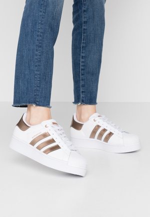 Białe sneakersy Adidas Superstar z metalicznymi brązowymi paskami i detalem na pięcie, noszone z krótkimi niebieskimi jeansami.