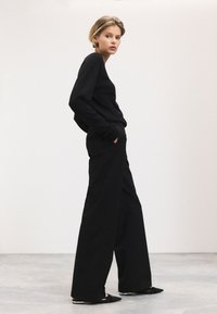 Haut noir à manches longues et pantalon à jambes larges, tous deux en tissu doux et texturé. Le modèle se tient les mains dans les poches, portant des chaussures plates noires.