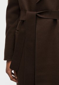 Σκούρο καφέ μάλλινο παλτό με φαρδύ ζώνη δεμένη γύρω από τη μέση, με μακριά μανίκια και ένα χέρι που ακουμπά κοντά στην πλαϊνή τσέπη.