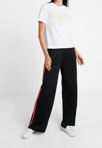 A woman stands in athletic wear, featuring a white t-shirt with a graphic and black wide-leg pants with red side stripes. She wears white sneakers.