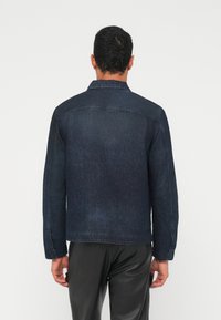 Tumši zila džinsa jaka ar apkakli, garām piedurknēm un taisnu griezumu. Aizmugures skats atklāj smalku izbalēšanu un šuvju detaļas.