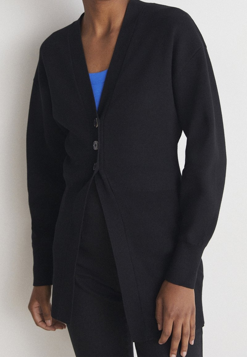 Personne portant un cardigan noir boutonné sur un haut bleu et un pantalon noir, debout devant un fond clair uni.