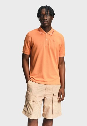 Uomo in piedi che indossa una polo arancione a maniche corte e pantaloncini cargo beige su uno sfondo grigio chiaro uniforme.