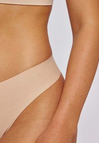 Naadloze, huidkleurige string met een soepele textuur en een brede tailleband. Huid die zichtbaar is aan de zijkant toont natuurlijke tinten, wat het comfort vergroot.