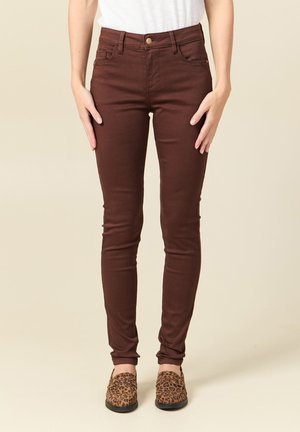 Kobieta ubrana w ciemnobrązowe obcisłe spodnie i wsuwane buty w panterkę, stojąca z rękami swobodnie opuszczonymi wzdłuż ciała.