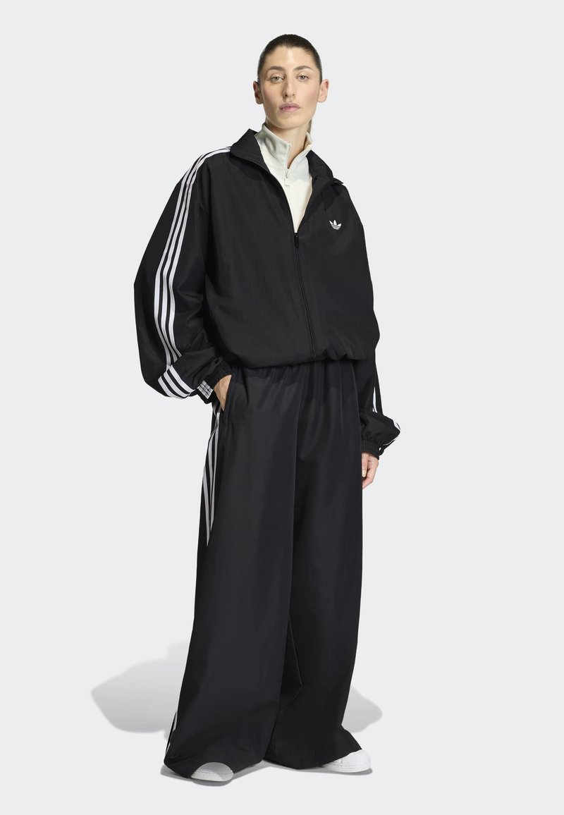 Donna che indossa un giubbotto sportivo Adidas nero e pantaloni sportivi a gamba larga con strisce bianche, in piedi con le mani nelle tasche contro uno sfondo semplice.