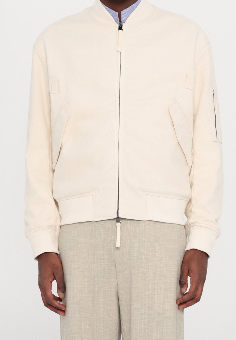 Veste bomber beige clair avec une fermeture éclair, des poches latérales et des poignets côtelés. Le tissu semble doux et légèrement texturé.