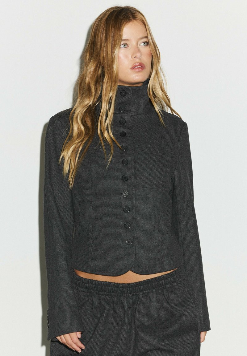 Femme blonde aux cheveux ondulés détachés porte une veste grise foncée boutonnée à col montant et un pantalon assorti sur un fond blanc uni.