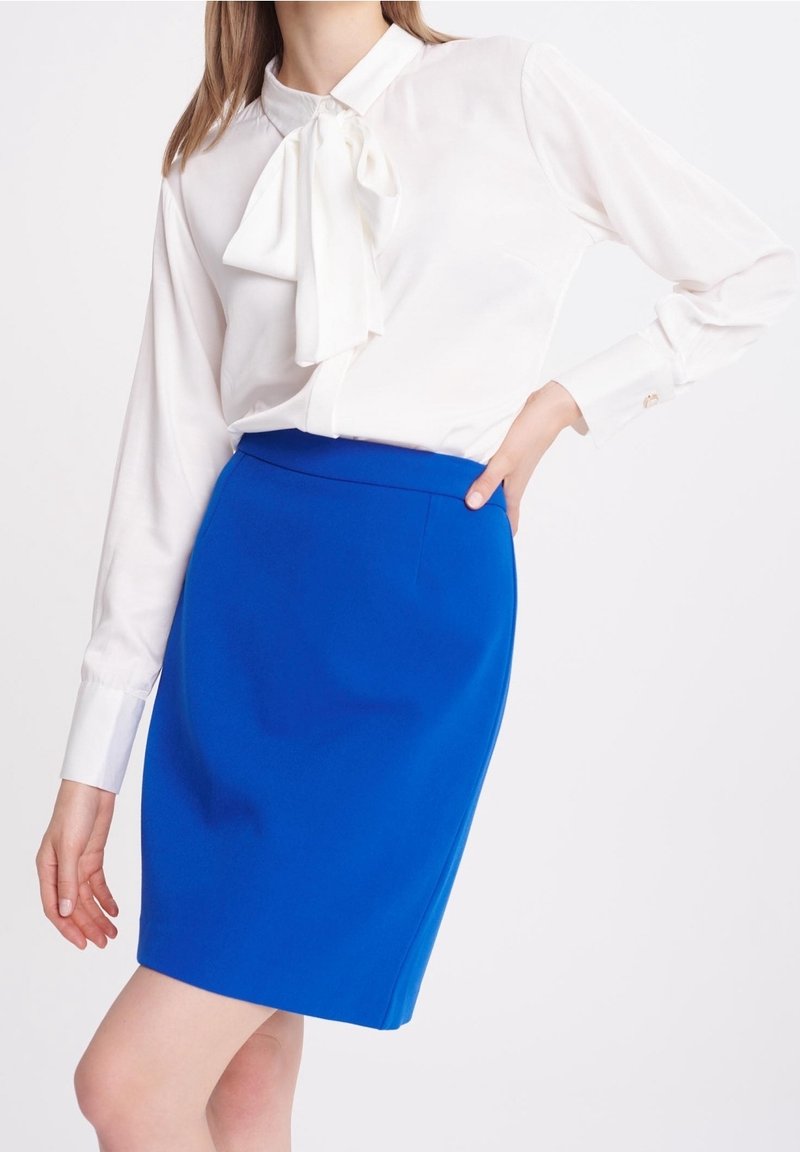 Bílá blůza s detaily na mašličku, kombinovaná s vykrojenou modrou tužkovou sukni. Hladká tkanina, upravený střih a jasný kontrast barev.