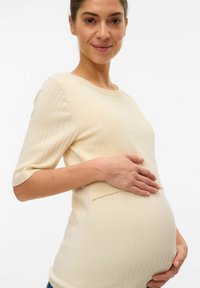 Maglietta premaman beige a coste con maniche corte, dotata di scollo rotondo e dettaglio della tasca, indossata su una pancia rotonda.