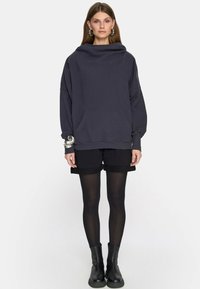 Kobieta ubrana w oversize'owy ciemnoszary hoodie, czarne szorty, przezroczyste czarne rajstopy, czarne botki za kostkę oraz duże srebrne kolczyki w kształcie koła, stojąca na tle białym.