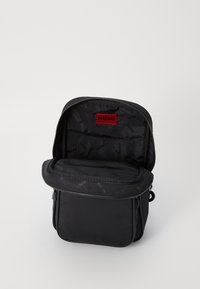 Sort nylon taske med lynlåslukning, der har en indvendig lomme og rød etikette. Blød tekstur, firkantet form og minimalistisk design.