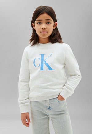 Bambino in piedi con una mano in tasca, che indossa una felpa bianca Calvin Klein e jeans azzurri chiari su uno sfondo grigio uniforme.