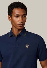 Granatowa koszulka polo z kontrastującym zielonym kołnierzykiem w paski, z zapięciem na guziki i haftowanym logo na piersi. Materiał wydaje się teksturowany.