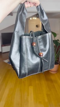 Črna usnjena torba tote z oblikovano strukturo, ki ima dekorativne bele šive in lesenimi poudarki na zapiranju. Ročaji so kratki in močni.