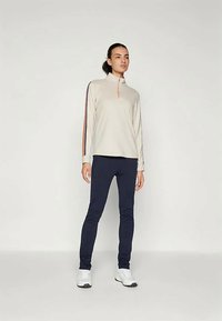 Woman standing wearing a beige long-sleeve top with striped sleeves, navy leggings, and white sneakers against a plain background.