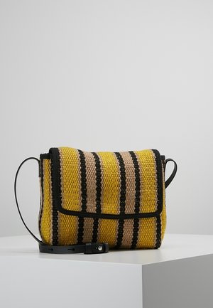 Żółta, beżowa i czarna paskowana pleciona torebka na ramię z czarną skórzaną lamówką i regulowanym paskiem na białej powierzchni.