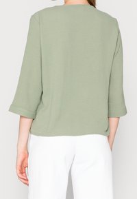 Blusa verde texturizzata con maniche a tre quarti, scollatura rotonda e vestibilità comoda, abbinata a pantaloni bianchi, vista dalla schiena.