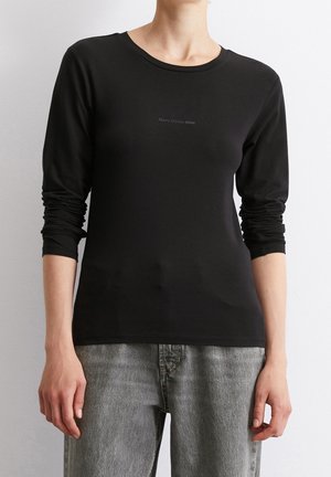 Persoon draagt een zwart aansluitend shirt met lange mouwen met het logo "Marc O’Polo DENIM" en grijze spijkerbroek, staand tegen een effen lichte achtergrond.