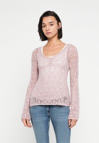 Ροζ πλεκτό πουλόβερ με περίπλοκο δαντελένιο σχέδιο, μακριά μανίκια, στρογγυλή λαιμόκοψη και λεπτομέρεια με κουμπιά στο πλάι, φορεμένο πάνω από λευκή μπλούζα.