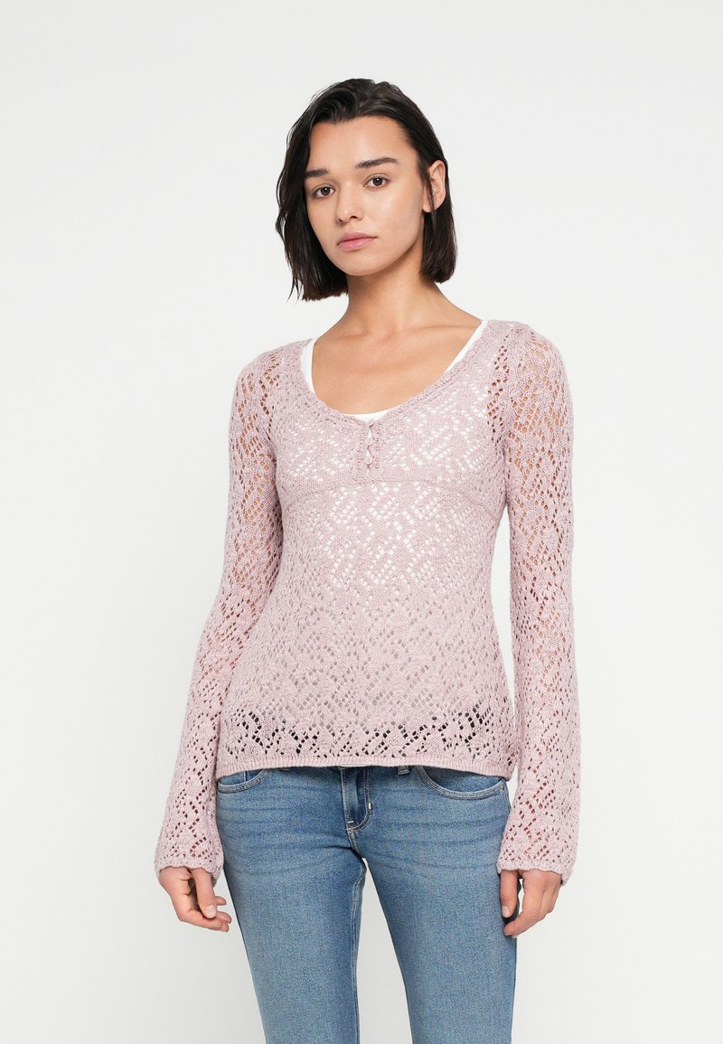 Ροζ πλεκτό πουλόβερ με περίπλοκο δαντελένιο σχέδιο, μακριά μανίκια, στρογγυλή λαιμόκοψη και λεπτομέρεια με κουμπιά στο πλάι, φορεμένο πάνω από λευκή μπλούζα.