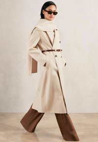 Beige wollenblend jas met een dubbelgebreide ontwerp, een ceintuur in de taille en een oversized kraag, gedragen met bruine wijde broek en een zonnebril.
