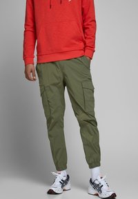 Olivenfärgade cargo byxor med elastiska muddar, flera fickor och en avslappnad passform; matchade med en röd hoodie och vita sneakers.