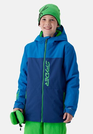 Enfant souriant portant une veste d'hiver bleue et marine avec une fermeture éclair verte et un lettrage, un bonnet vert avec motif d'araignée, et un pantalon vert tenant des gants verts.