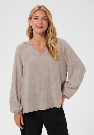 Femme blonde portant un chemisier beige ample à manches longues avec une texture florale subtile et un pantalon noir, souriante, une main sur la hanche.