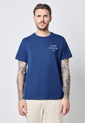 T-shirt en coton bleu marine à manches courtes, avec un texte blanc qui dit "LE SOLEIL, TOUTE L'ANNÉE" sur la partie supérieure gauche de la poitrine.