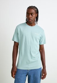 Lyseblå bomulds T-shirt med korte ærmer, rund hals og et lille guld logo på brystet. Sammen med blå denim jeans.