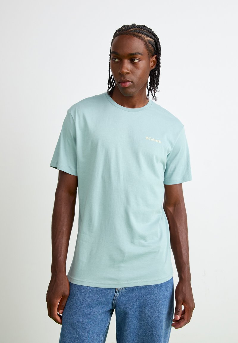 T-shirt en coton bleu clair avec des manches courtes, col rond et un petit logo doré sur la poitrine. Associé à un jean en denim bleu.