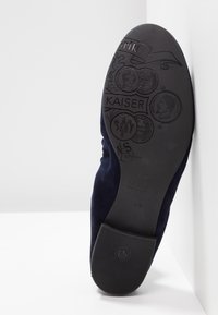 Semelle d'une chaussure noire montrant des motifs en relief, incluant des pièces, le mot Kaiser et la taille 41, inclinée contre un mur d'angle blanc.
