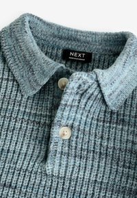 Texturerad blå stickad tröja med ribbat mönster, har en krage, två knappar och en etikett som säger "NEXT," tillverkad i Myanmar.