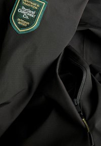 Černá látková bunda s zelenou a zlatou nášivkou "Portland Outerwear Co." Podél švu je viditelný detail se zipovou kapsou.