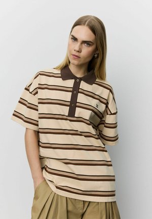 Joven con cabello liso castaño claro que lleva una camiseta polo beige y marrón de manga corta a rayas y pantalones beige, de pie contra una pared blanca.