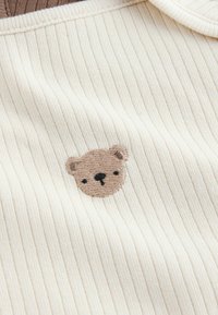 Cream striped fabric with a ribbed texture featuring a small, embroidered bear face in muted brown tones, centered on the material.